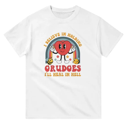 White T-shirt with a playful cartoon design of a red heart character flashing peace signs, surrounded by a rainbow arch and yellow smiley flowers. The shirt features bold text reading, "I BELIEVE IN HOLDING GRUDGES, I'LL HEAL IN HELL," in a retro-style font, blending humor with a touch of sarcasm.