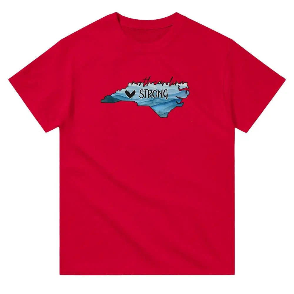 Red T-shirt featuring an outline of North Carolina filled with blue and green tones, symbolizing a resilient landscape. Above the shape, the text reads "North Carolina" in a cursive font, with "STRONG" written below alongside a small heart icon. This design conveys a message of support and resilience, possibly for hurricane relief efforts.