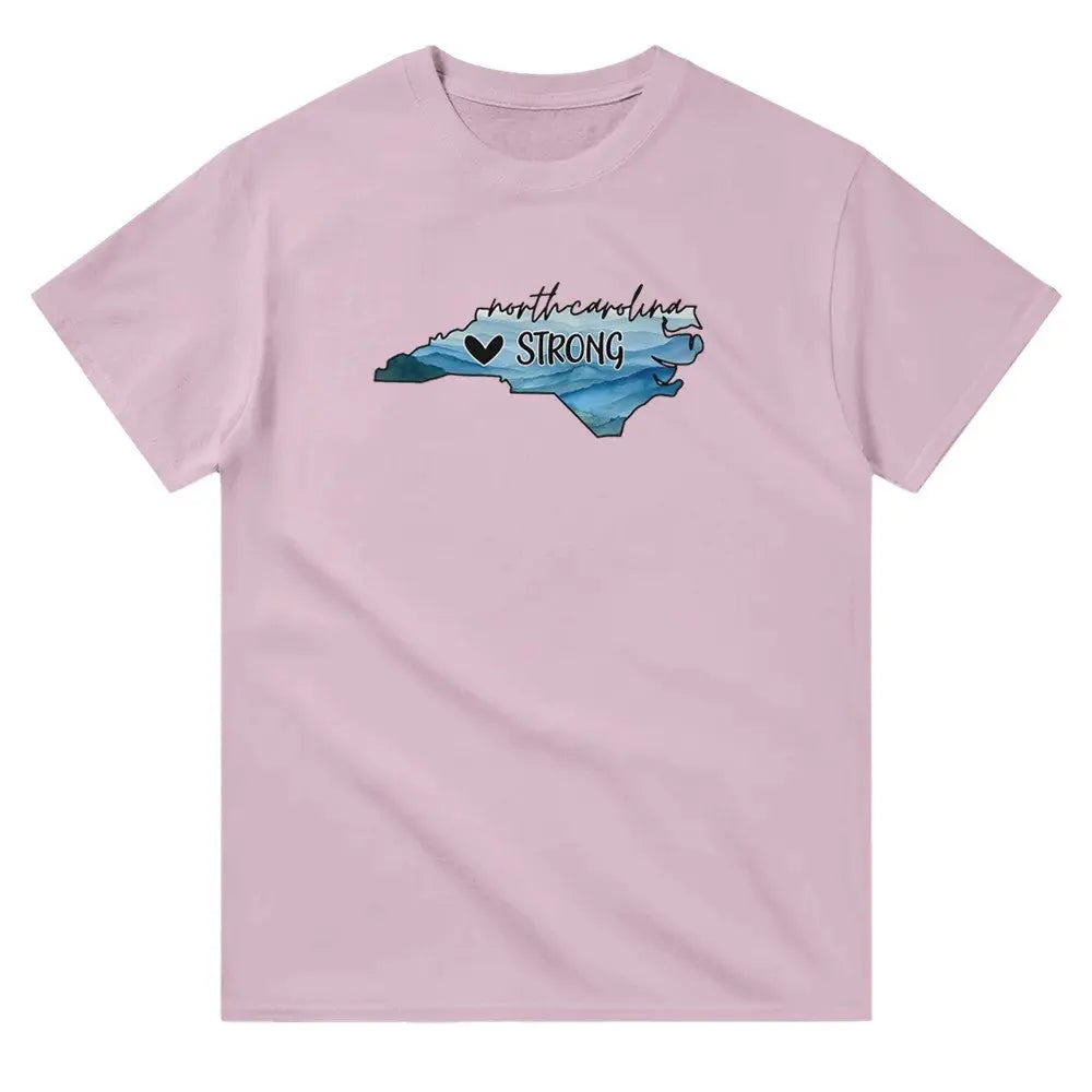 Pink T-shirt featuring an outline of North Carolina filled with blue and green tones, symbolizing a resilient landscape. Above the shape, the text reads "North Carolina" in a cursive font, with "STRONG" written below alongside a small heart icon. This design conveys a message of support and resilience, possibly for hurricane relief efforts.