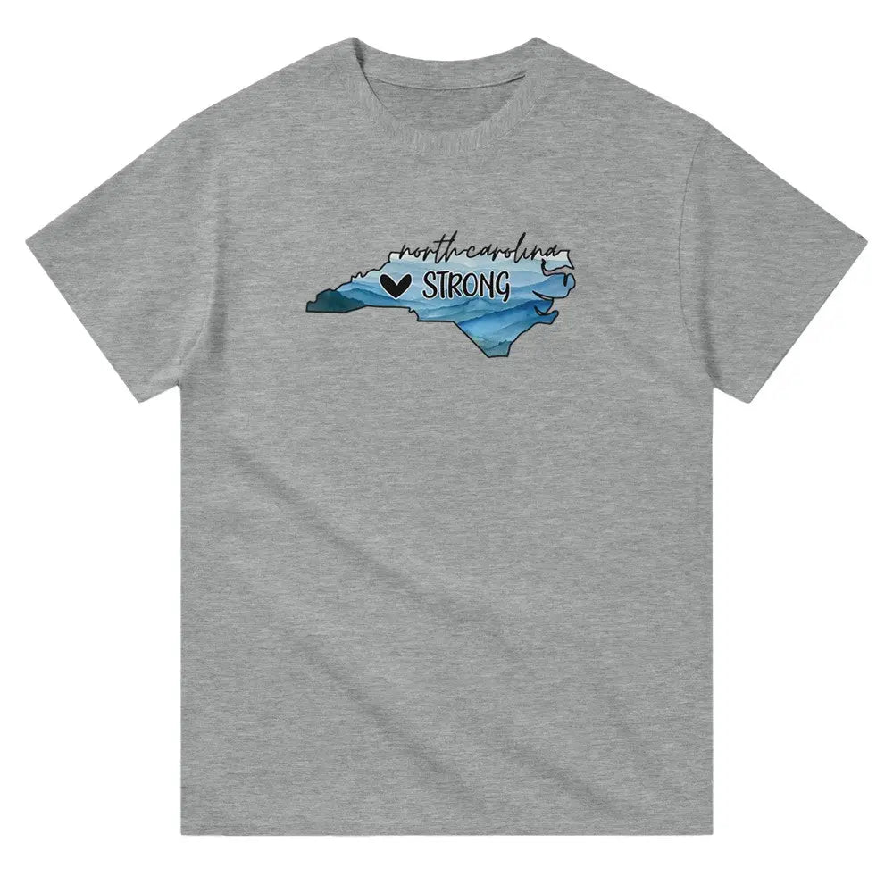 Grey T-shirt featuring an outline of North Carolina filled with blue and green tones, symbolizing a resilient landscape. Above the shape, the text reads "North Carolina" in a cursive font, with "STRONG" written below alongside a small heart icon. This design conveys a message of support and resilience, possibly for hurricane relief efforts.