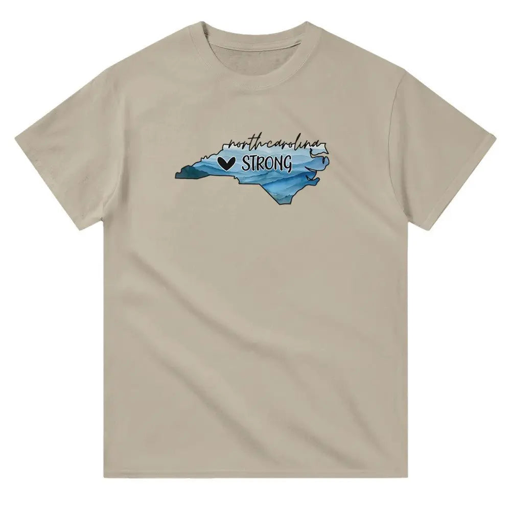 Beige T-shirt featuring an outline of North Carolina filled with blue and green tones, symbolizing a resilient landscape. Above the shape, the text reads "North Carolina" in a cursive font, with "STRONG" written below alongside a small heart icon. This design conveys a message of support and resilience, possibly for hurricane relief efforts.