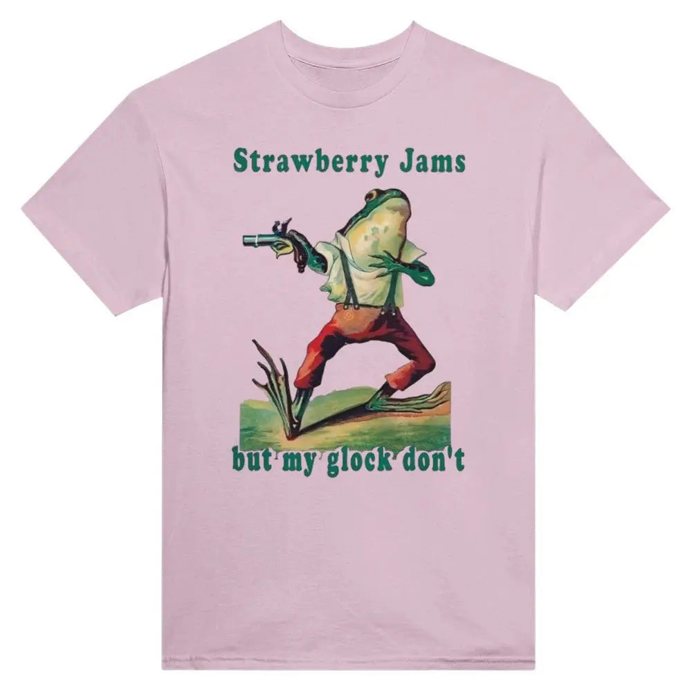 Pink T-shirt featuring a humorous illustration of a frog dressed in vintage-style clothing, holding a gun. Above the frog, the text reads, "Strawberry Jams, but my glock don't," in a playful, retro font. The design combines whimsical art with a tongue-in-cheek message.