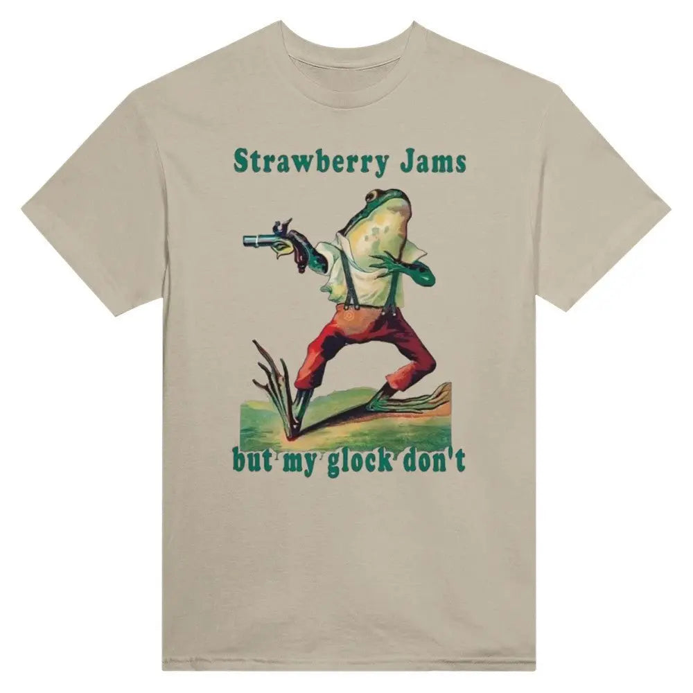 Beige T-shirt featuring a humorous illustration of a frog dressed in vintage-style clothing, holding a gun. Above the frog, the text reads, "Strawberry Jams, but my glock don't," in a playful, retro font. The design combines whimsical art with a tongue-in-cheek message.