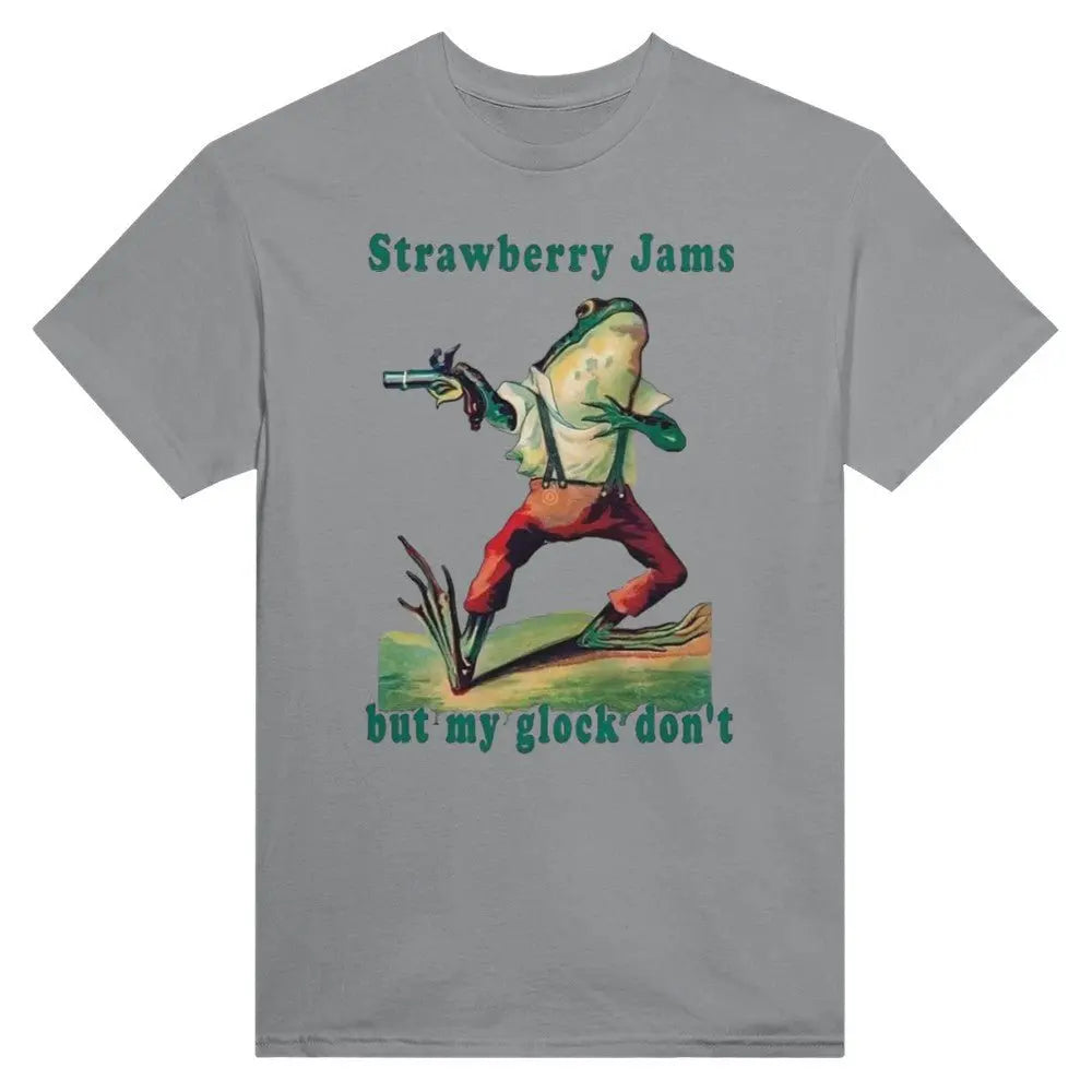 Grey T-shirt featuring a humorous illustration of a frog dressed in vintage-style clothing, holding a gun. Above the frog, the text reads, "Strawberry Jams, but my glock don't," in a playful, retro font. The design combines whimsical art with a tongue-in-cheek message.